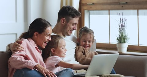 Cute small child girl using educational application on computer, sitting on couch with smaller sister and smiling parents. Happy parents enjoying fun time, watching cartoons video movies with kids.