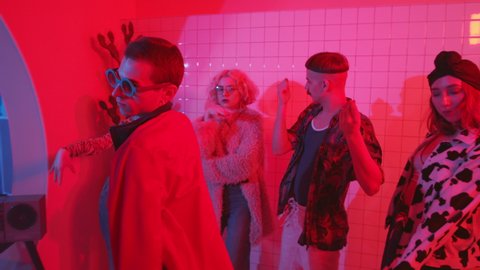 Young eccentric man in trendy wear and sunglasses dancing expressive vogue with group of stylish friends in room with color neon light and fog