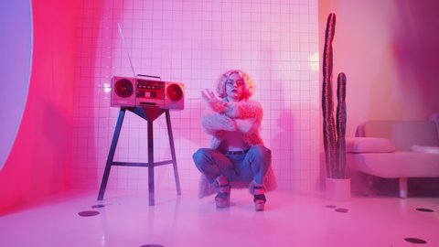 Young blonde female dancer in glamorous outfit showing vogue hands performance and looking at camera while sitting in squat near retro cassette player in studio with pink neon light
