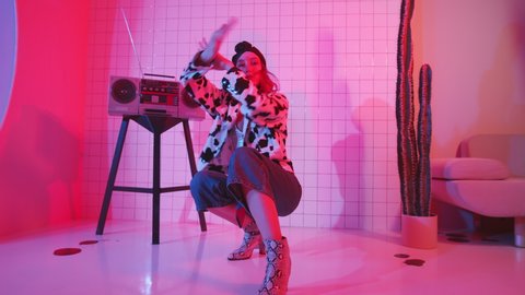 Professional female dancer in trendy outfit and high heels dancing vogue on the floor for camera in studio with pink neon light and vintage cassette player