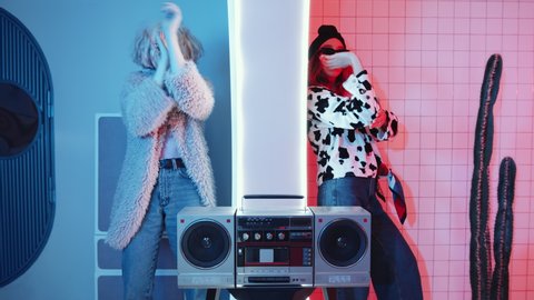 Two young women in glamorous outfits dancing femme vogue on both sides of the wall between different rooms with pink and blue neon lighting and retro cassette player
