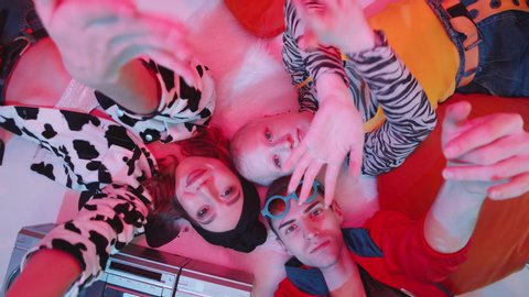 Top view shot of three young dancers in stylish outfits lying on the floor with retro cassette player, posing for camera and performing vogue hands movements under pink lighting