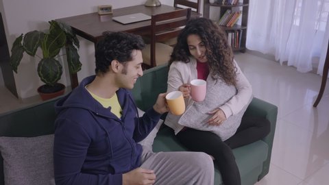 A Smiling happy Husband offers a hot cup of coffee to a beautiful wife on a cold winter day. Attractive man male and woman or female sitting on a couch enjoying a steamy cup of tea on a cozy morning.