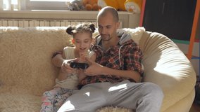Dad and daughter play video games together sitting on the sofa in a Sunny room.
