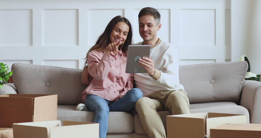 Happy young family couple sitting on sofa among huge carton boxes with belongings, using computer digital tablet, buying purchasing decoration items online for new apartment home, moving day concept. Royalty-Free Stock Footage #1049938138