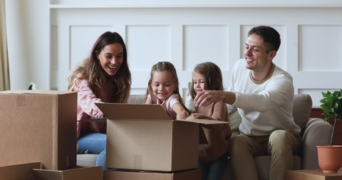 Playful family smiling little adorable kids sisters helping happy young couple parents unpacking belongings from huge cardboard boxes, sitting on sofa in new apartment home, moving day concept.