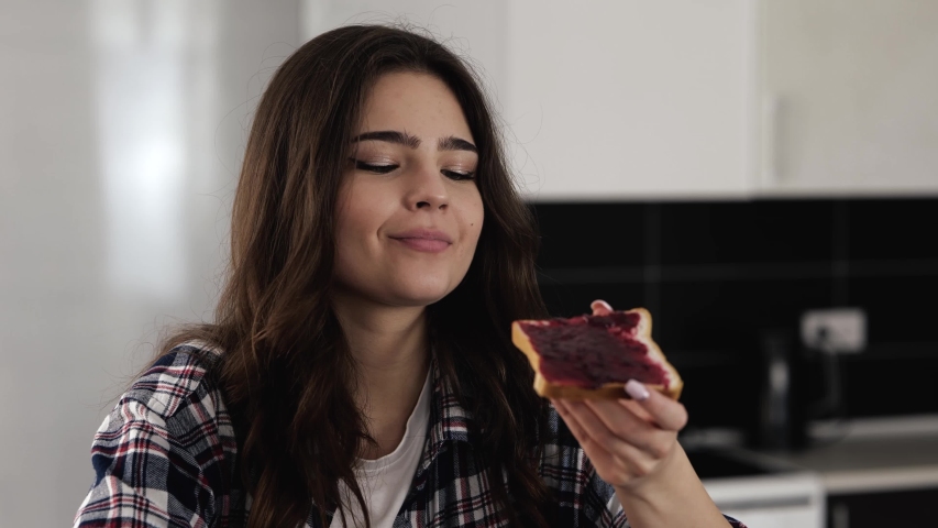 Young woman in kitchen during quarantine. Sit and eat tasty delicious piece of bread with jam. Enjoying meal breakfast in morning. | Shutterstock HD Video #1049940022