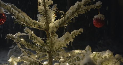 Snowy Christmas tree with lights and decorations outside in forest while snowing