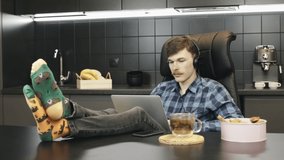 Young male in headphones listening to music and working on laptop computer from home. Man relaxing and working at home office. Guy using notebook sitting in kitchen at home workplace