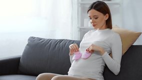 Laughing pregnant girl holding pink baby booties on couch