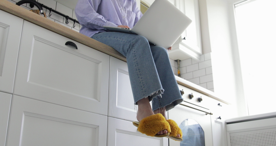 Young attractive casual woman is sitting in kitchen workplace desk and working on laptop drinking coffee. Wear cute yellow fluffy domestic slippers and blue jeans with shirt | Shutterstock HD Video #1049953792