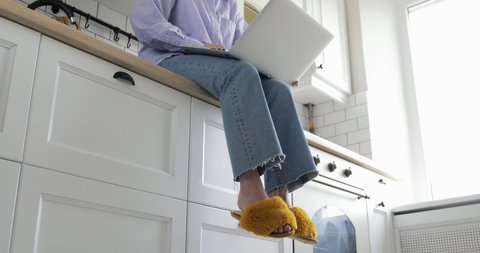 Young attractive casual woman is sitting in kitchen workplace desk and working on laptop drinking coffee. Wear cute yellow fluffy domestic slippers and blue jeans with shirt