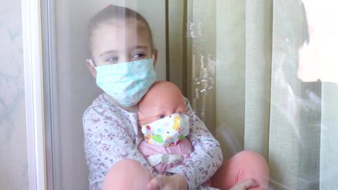 Quarantine, threat of coronavirus. Sad child and his doll both in protective medical masks sits on windowsill and looks out window. Virus protection, pandemic, prevention epidemic.