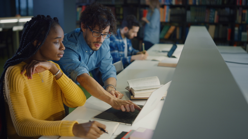 University Library: Gifted Black Girl uses Laptop, Smart Classmate Explains and Helps Her with Class Assignment. Happy Diverse Students Talking, Learning, Studying Together for Exams Royalty-Free Stock Footage #1049967724