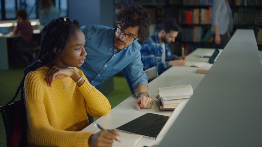 University Library: Gifted Black Girl uses Laptop, Smart Classmate Explains and Helps Her with Class Assignment. Happy Diverse Students Talking, Learning, Studying Together for Exams Royalty-Free Stock Footage #1049967724