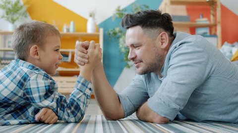 Middle-aged man and little boy are practising arm wresting at home enjoying activity, child is winning. Modern pastime, family relationship and fun concept.