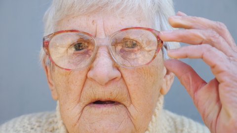 Portrait of old lady in eyeglasses with pensive sight. Close up wrinkled face of female pensioner looking into camera and adjusting glasses. Sorrow facial expression of granny. Gaze of mature woman
