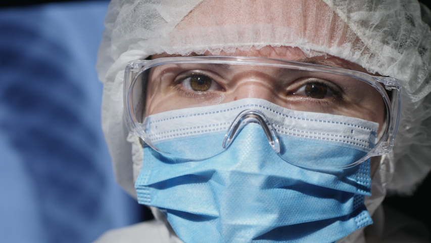 Close-up face of a woman in medical protective uniform, goggles and a face mask. Medic looks at the camera, working in the clinic during an epidemic. Royalty-Free Stock Footage #1049974639