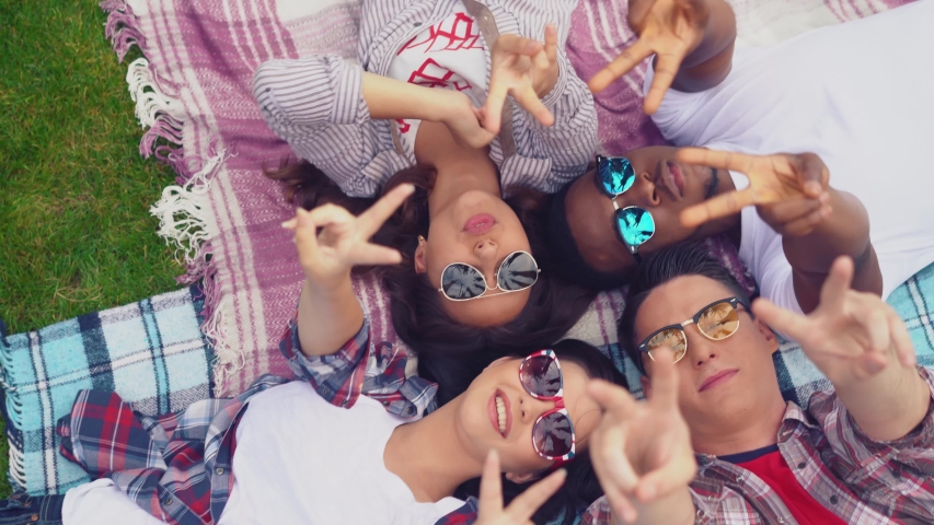 Young carefree mixed race students laying on a plaid on a green grass at campus park, enjoying free summer time together and shoving gesture or a hand sign victory tuting hands together making a star