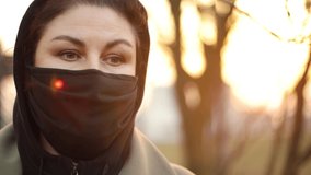 Video footage slow motion, a young adult woman in a black protective mask on her face walks through the street on a spring day at sunset in sunny glare.