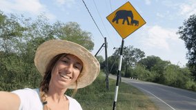Young woman taking selfie on the side of the road near elephants crossing sign in Sri Lanka national park. Girl video chatting selfie while enjoying road trip adventures in Asia 