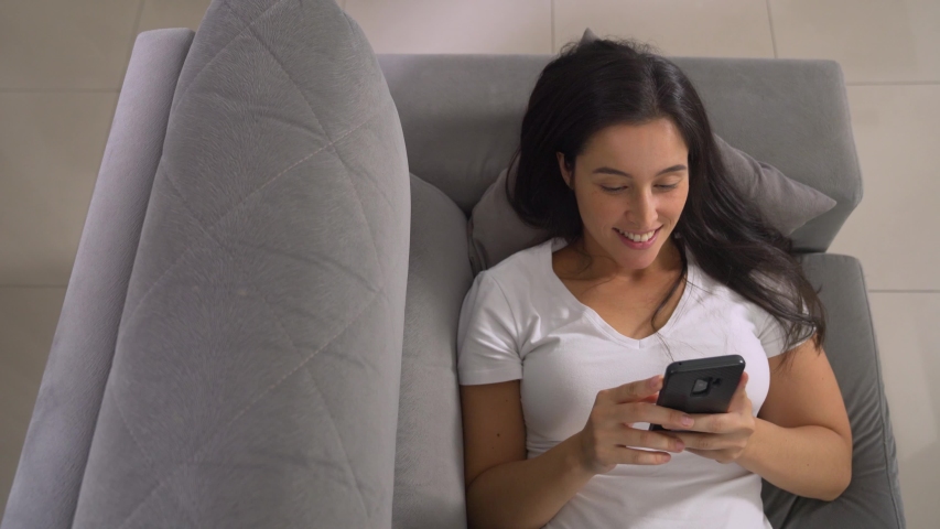 Happy relaxed girl holding smart phone using mobile apps watching funny video laughing lying on couch, smiling lazy young woman having fun chatting in social media resting on sofa at home, top view
 | Shutterstock HD Video #1049981842