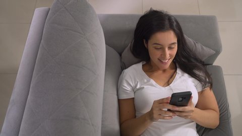 Happy relaxed girl holding smart phone using mobile apps watching funny video laughing lying on couch, smiling lazy young woman having fun chatting in social media resting on sofa at home, top view
