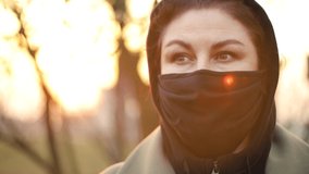 Video footage, a young adult woman in a black protective mask on her face walks through the street on a spring day at sunset in sunny glare.
