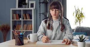 Portrait of cute little girl with braided hairstyle sitting at home and painting pictures. Teenager in domestic clothing enjoying leisure time with art.