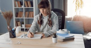 Pretty young lady with trendy hairstyle sitting at table and studying art. Teenager in grey hoodie doing homework while staying at home.