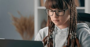 Serious schoolgirl with trendy braids learning lessons online while sitting at home with laptop. Charming teenager in eyeglasses doing homework during quarantine time.