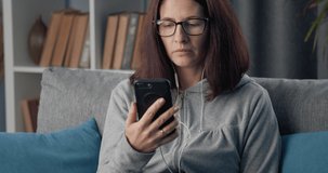 Serious mature woman in eyeglasses having online conversation on smartphone while staying at home. Female freelancer working on comfortable grey couch.