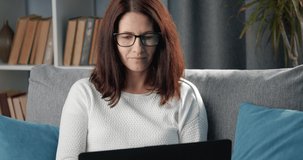 Portrait of concentrated woman in eyewear and white sweater sitting on cozy sofa and working on personal laptop. Concept of of remote work and female career.