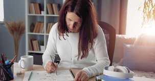 Beautiful woman in white sweater sitting at home and drawing pictures. Mature female with dark hair painting with colorful pencils. Concept of creativity.