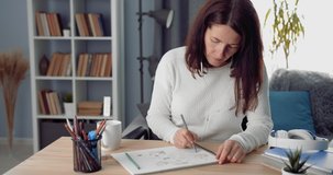 Mature lady with dark hair sitting at table and drawing with colored pencils in album. Beautiful woman in casual clothing entertaining her self during free time at home.