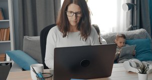 Confident mature lady in eyewear and white sweater working on laptop while her two children sitting behind and studying at home. Concept of quarantine lifestyle.
