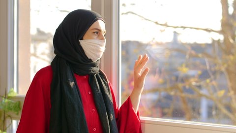 Self isolation. Woman with hijab and wear mask looks out of house window depressed and bored. Arab woman lokking out window. Mental health,self isolation in own home, corona virus covid-19. 