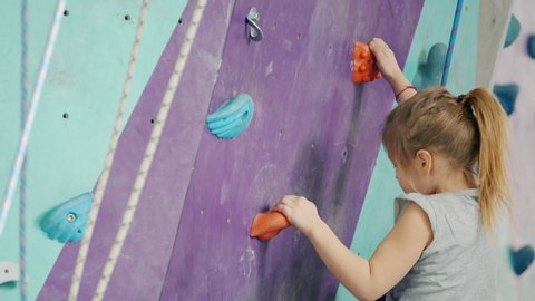 Brave child little girl is climbing up artificial wall in modern rock-climbing gym exercising alone using safety equipment. Childhood and extreme sports concept.