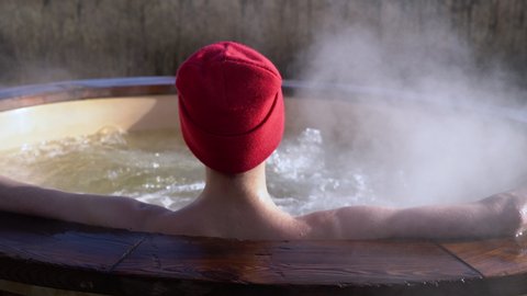 Young adult relaxing in wooden hot tub outside and looking at nature. Person enjoying hot steaming pool on a sunny day, private spa treatment
