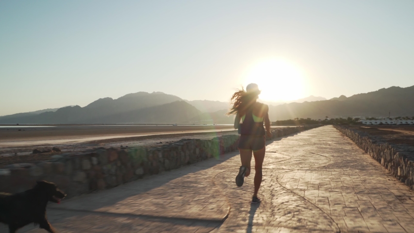 Rear view female runs with her funny dog in dry desert pavement road at sunrise