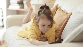 Little Girl in Yellow Hoodie Lying on Sofa and Holding Smartphone in her Hands, Playing Mobile Video Game. Sunny Yellow Cozy Home Interior.