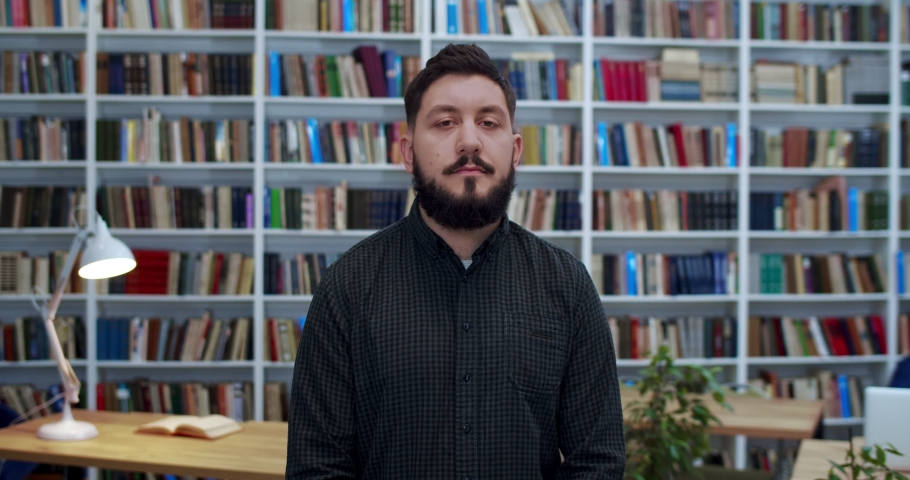 Portrait of young Caucasian man with beard standing in libary, crossing hands in front of him and looking at camera. Male professor or worker of bibliotheca. Books shelves on background. Wise teacher.