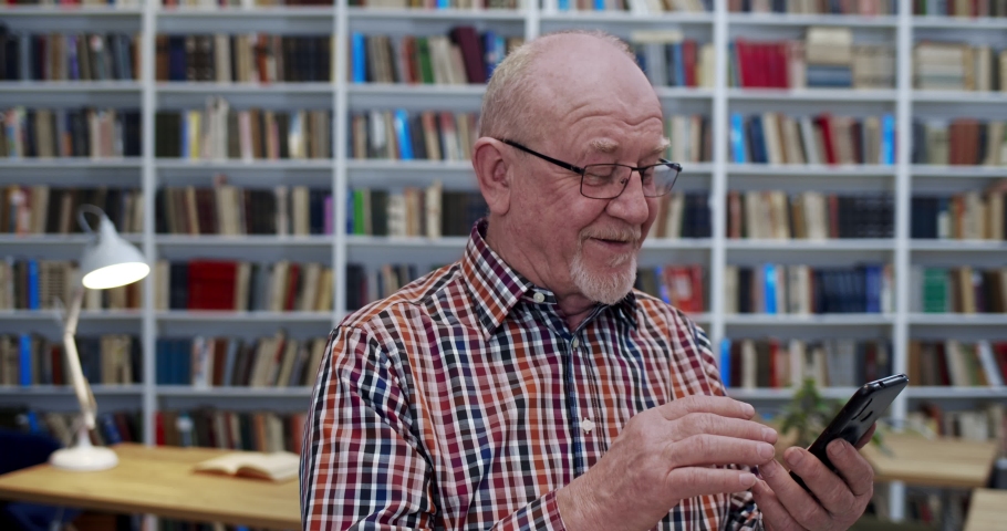 Old Cucasian cheerful bald male library worker in glasses and plaid shirt tapping and texting message on mobile phone. Happy man senior professor typing and scrolling on smartphone in bibliotheca.