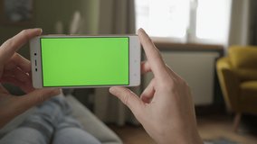 Young Woman Home Lying on a Couch with Green Screen Smartphone in Horizontal Mode. Girl Using Touchscreen Mobile Phone. Girl Using Smartphone, Browsing Internet, Watching Video Content, Blogs. POV.