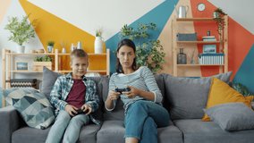 Little child is playing video games with caring mother holding joysticks pushing buttons and talking smiling at home. Modern devices and happy youth concept.