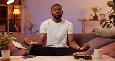 Young handsome man doing breathing exercise while sitting on sofa at home. Bearded african guy doing yoga while relaxing at free time . Concept of healthy lifestyle and leisure.