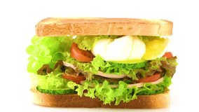 Sandwich with fresh lettuce, ham, cucumber, tomato and poached eggs isolated on white background. Boiled egg is cut and a liquid yolk flows from it. Close-up. Selective focus. 4K UHD video 3840x2160