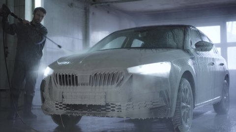 Car with turned on headlights standing in car wash service. Worker washing off foam from vehicle surface with high-pressure sprayer. Maintenance, industry, business.