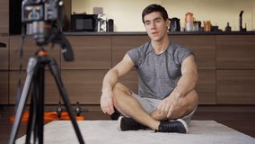 Young fitness influencer sitting cross-legged on floor at home and speaking at camera while filming video blog