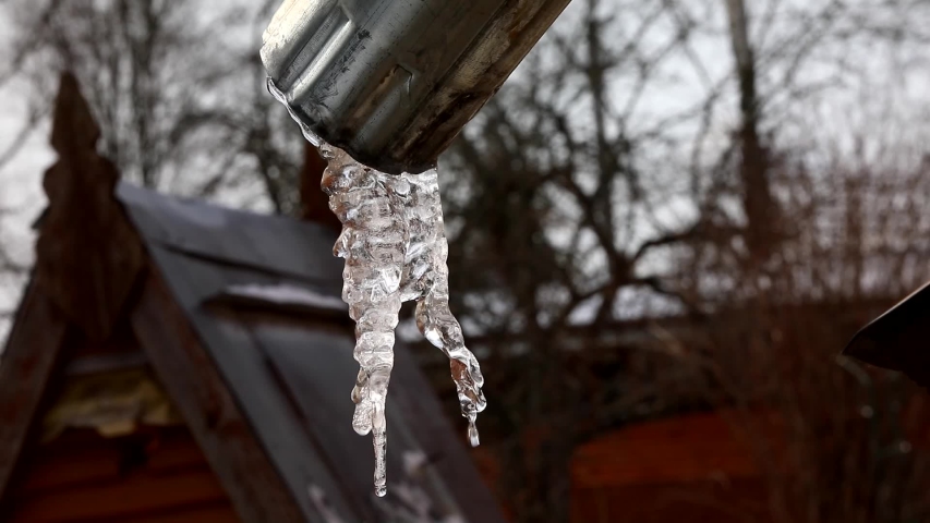 A large transparent icicle of a bizarre shape in the drainpipe drips profusely on the blurred background of the patio of a country house on a wet day.Spring drops.The final melting ice.Russia